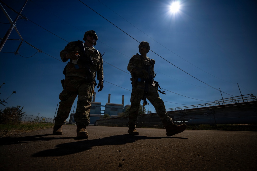 Two armed military or security personnel in full combat gear are walking along a paved road in a silhouetted low-angle shot against a bright blue sky. The image is taken from a dramatic low perspective with the sun creating a strong backlight effect. An industrial facility with smokestacks is visible in the background.
