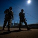 Two armed military or security personnel in full combat gear are walking along a paved road in a silhouetted low-angle shot against a bright blue sky. The image is taken from a dramatic low perspective with the sun creating a strong backlight effect. An industrial facility with smokestacks is visible in the background.