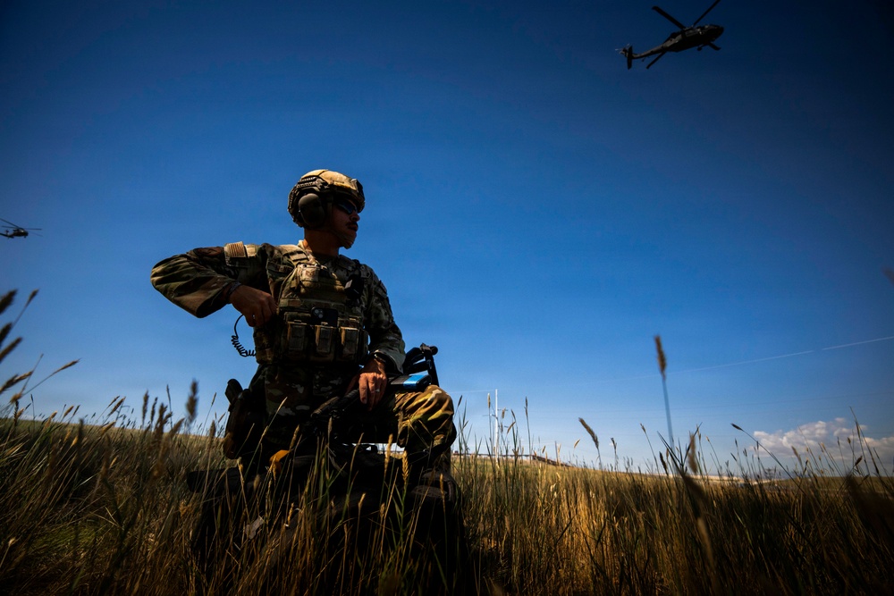 A soldier in full tactical gear stands in a field of tall dry grass under a clear blue sky, holding what appears to be a tablet or communication device. At least two military helicopters are visible in the background sky, suggesting an active field operation or training exercise. The low-angle shot creates a dramatic silhouette effect against the bright sky.