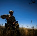 A soldier in full tactical gear stands in a field of tall dry grass under a clear blue sky, holding what appears to be a tablet or communication device. At least two military helicopters are visible in the background sky, suggesting an active field operation or training exercise. The low-angle shot creates a dramatic silhouette effect against the bright sky.
