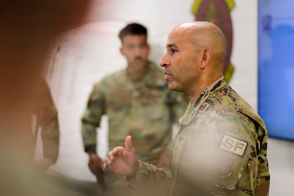 Two military personnel in OCP (Operational Camouflage Pattern) uniforms are present in what appears to be a briefing or meeting room. The foreground individual, wearing an 'SF' (Security Forces) patch, appears to be speaking or presenting while gesturing with his hands. A unit insignia/patch is visible on the wall in the background.
