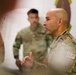 Two military personnel in OCP (Operational Camouflage Pattern) uniforms are present in what appears to be a briefing or meeting room. The foreground individual, wearing an 'SF' (Security Forces) patch, appears to be speaking or presenting while gesturing with his hands. A unit insignia/patch is visible on the wall in the background.