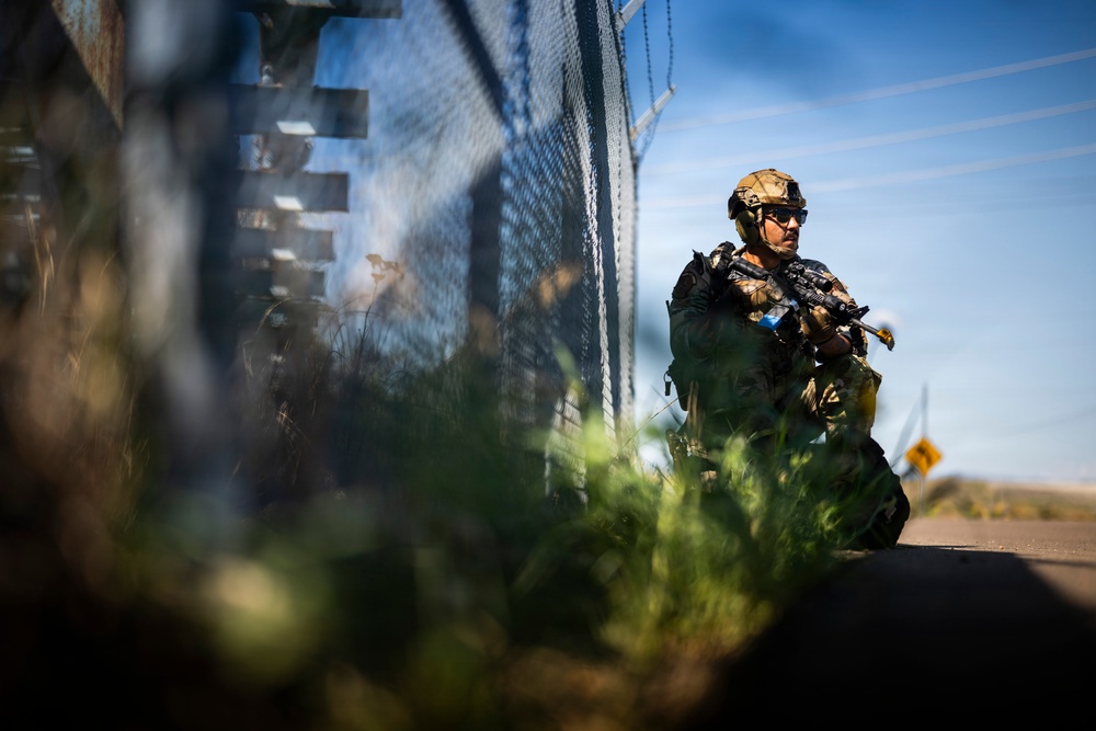 A heavily armed and equipped soldier or tactical operator is kneeling in a low-profile position alongside a tall chain-link or mesh security fence. The individual is wearing full tactical gear including a ballistic helmet, body armor, and carrying a rifle. The image is shot from a low ground-level perspective with foreground vegetation creating a shallow depth-of-field effect.