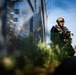A heavily armed and equipped soldier or tactical operator is kneeling in a low-profile position alongside a tall chain-link or mesh security fence. The individual is wearing full tactical gear including a ballistic helmet, body armor, and carrying a rifle. The image is shot from a low ground-level perspective with foreground vegetation creating a shallow depth-of-field effect.