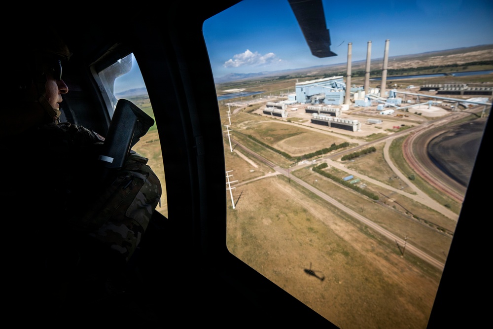 A person in military camouflage uniform is seated inside a helicopter, looking out a large window at an industrial facility below. The aerial view reveals what appears to be a coal-fired power plant or similar heavy industrial complex with tall smokestacks, large buildings, and surrounding infrastructure. The helicopter's shadow is visible on the ground below.