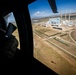 A person in military camouflage uniform is seated inside a helicopter, looking out a large window at an industrial facility below. The aerial view reveals what appears to be a coal-fired power plant or similar heavy industrial complex with tall smokestacks, large buildings, and surrounding infrastructure. The helicopter's shadow is visible on the ground below.