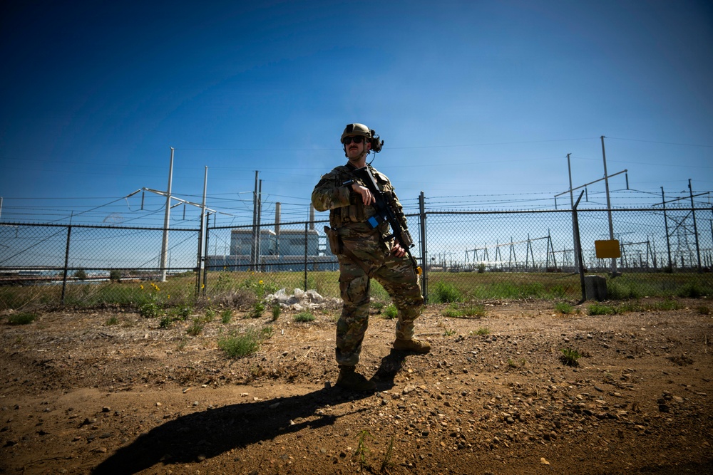 A fully equipped military or law enforcement operator stands in a security posture outside a fenced industrial or utility facility. The individual is wearing multicam camouflage, tactical gear, and a helmet with mounted equipment, and is carrying a rifle. The setting appears to be a perimeter security operation at what looks like a power generation or electrical substation facility.