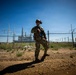 A fully equipped military or law enforcement operator stands in a security posture outside a fenced industrial or utility facility. The individual is wearing multicam camouflage, tactical gear, and a helmet with mounted equipment, and is carrying a rifle. The setting appears to be a perimeter security operation at what looks like a power generation or electrical substation facility.