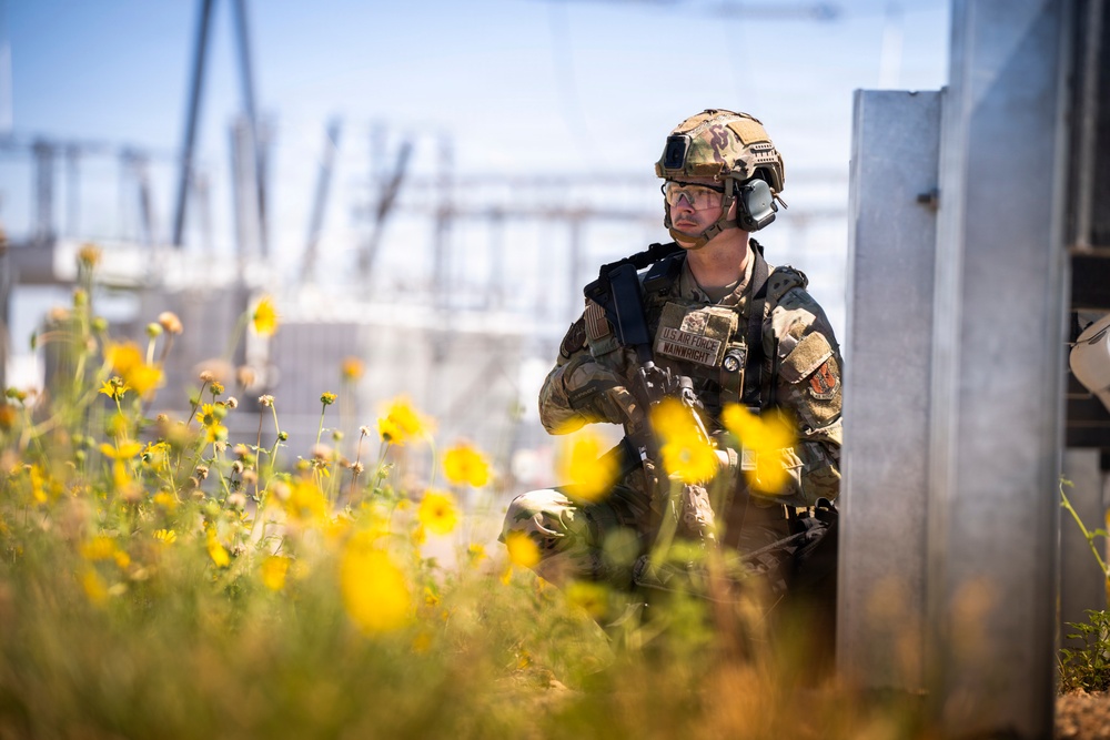A U.S. Air Force service member in full tactical gear is positioned in a kneeling stance beside a metal structure, appearing to conduct a security or reconnaissance exercise near what appears to be an electrical substation. The soldier's name tape reads 'WAINWRIGHT' and the branch tape reads 'U.S. AIR FORCE.' The foreground is filled with yellow wildflowers, creating a striking visual contrast with the military subject.