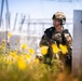 A U.S. Air Force service member in full tactical gear is positioned in a kneeling stance beside a metal structure, appearing to conduct a security or reconnaissance exercise near what appears to be an electrical substation. The soldier's name tape reads 'WAINWRIGHT' and the branch tape reads 'U.S. AIR FORCE.' The foreground is filled with yellow wildflowers, creating a striking visual contrast with the military subject.