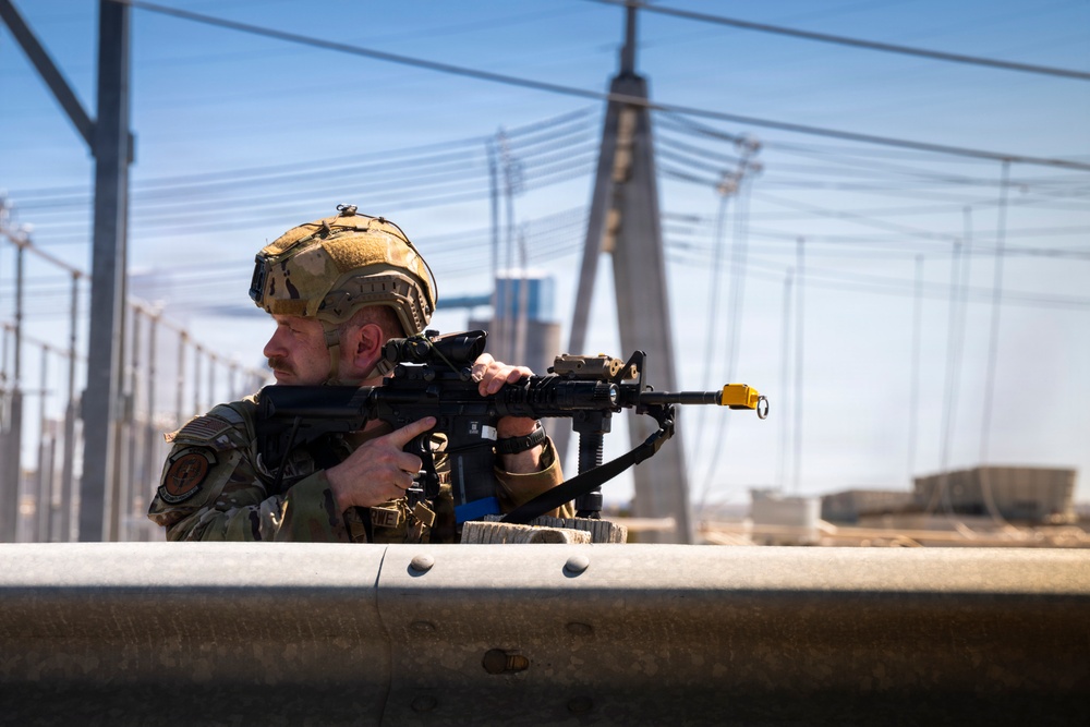A soldier in full tactical gear is positioned behind a concrete barrier, aiming a rifle with optical attachments. The setting appears to be an urban or industrial environment, with a cable-stayed bridge and power infrastructure visible in the background. The image is a high-quality, sharp photograph likely taken during a military operation or training exercise.