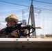 A soldier in full tactical gear is positioned behind a concrete barrier, aiming a rifle with optical attachments. The setting appears to be an urban or industrial environment, with a cable-stayed bridge and power infrastructure visible in the background. The image is a high-quality, sharp photograph likely taken during a military operation or training exercise.