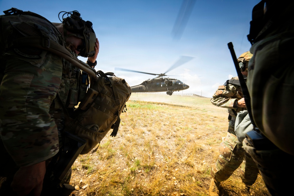 Two military personnel in full combat gear are crouching or bracing against rotor wash as a UH-60 Black Hawk helicopter hovers or lands nearby on an open grassy field. The soldiers are wearing multicam/OCP camouflage uniforms, tactical helmets, and carrying large rucksacks and weapons. The scene appears to depict a helicopter insertion or extraction operation during a military exercise or operation.
