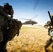 Two military personnel in full combat gear are crouching or bracing against rotor wash as a UH-60 Black Hawk helicopter hovers or lands nearby on an open grassy field. The soldiers are wearing multicam/OCP camouflage uniforms, tactical helmets, and carrying large rucksacks and weapons. The scene appears to depict a helicopter insertion or extraction operation during a military exercise or operation.