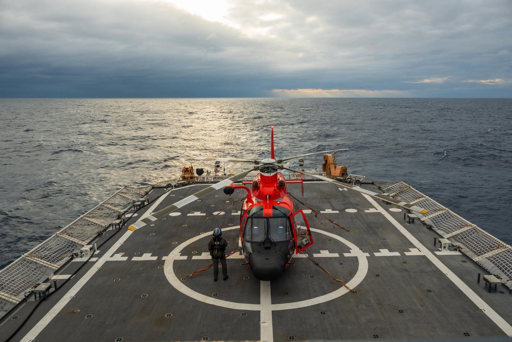 A red and gray helicopter, consistent with a U.S. Coast Guard MH-65 Dolphin, is secured on the flight deck of a Coast Guard cutter at sea. A single crew member in flight gear stands near the aircraft on the marked helipad. The scene is set under overcast skies with calm ocean waters visible on both sides of the vessel.