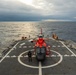 A red and gray helicopter, consistent with a U.S. Coast Guard MH-65 Dolphin, is secured on the flight deck of a Coast Guard cutter at sea. A single crew member in flight gear stands near the aircraft on the marked helipad. The scene is set under overcast skies with calm ocean waters visible on both sides of the vessel.