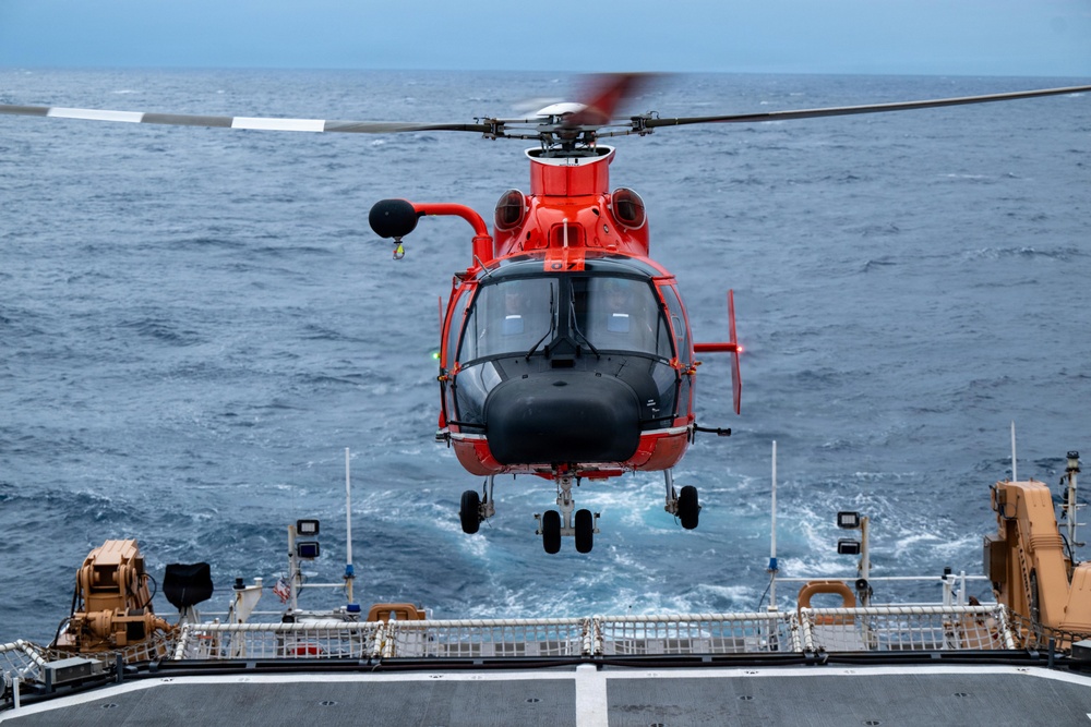 A red and black helicopter, consistent with a U.S. Coast Guard MH-65 Dolphin, is hovering just above the flight deck of a naval or coast guard vessel at sea. The helicopter appears to be in the process of landing or departing from the ship's stern flight deck. The ocean is visible in the background with choppy blue-gray water under overcast skies.