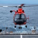 A red and black helicopter, consistent with a U.S. Coast Guard MH-65 Dolphin, is hovering just above the flight deck of a naval or coast guard vessel at sea. The helicopter appears to be in the process of landing or departing from the ship's stern flight deck. The ocean is visible in the background with choppy blue-gray water under overcast skies.
