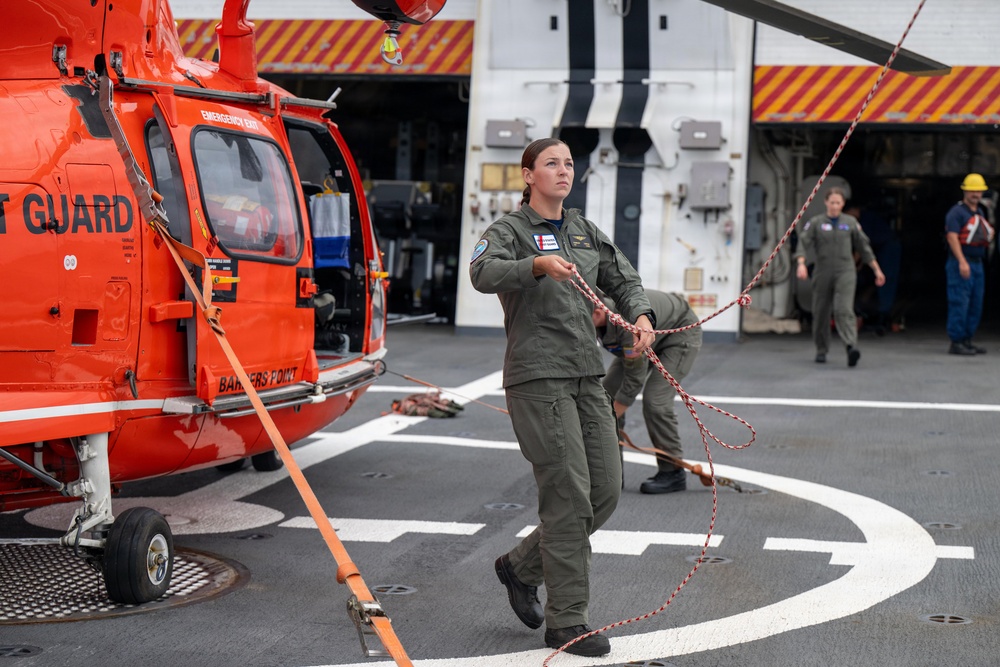 A U.S. Coast Guard crew is working on a ship's flight deck with an orange Coast Guard helicopter labeled 'BARBERS POINT.' Personnel in olive/sage green flight suits are handling tie-down ropes and securing the aircraft on what appears to be a vessel's helipad. Additional crew members in flight suits and safety gear are visible in the background near a hangar bay.