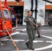 A U.S. Coast Guard crew is working on a ship's flight deck with an orange Coast Guard helicopter labeled 'BARBERS POINT.' Personnel in olive/sage green flight suits are handling tie-down ropes and securing the aircraft on what appears to be a vessel's helipad. Additional crew members in flight suits and safety gear are visible in the background near a hangar bay.