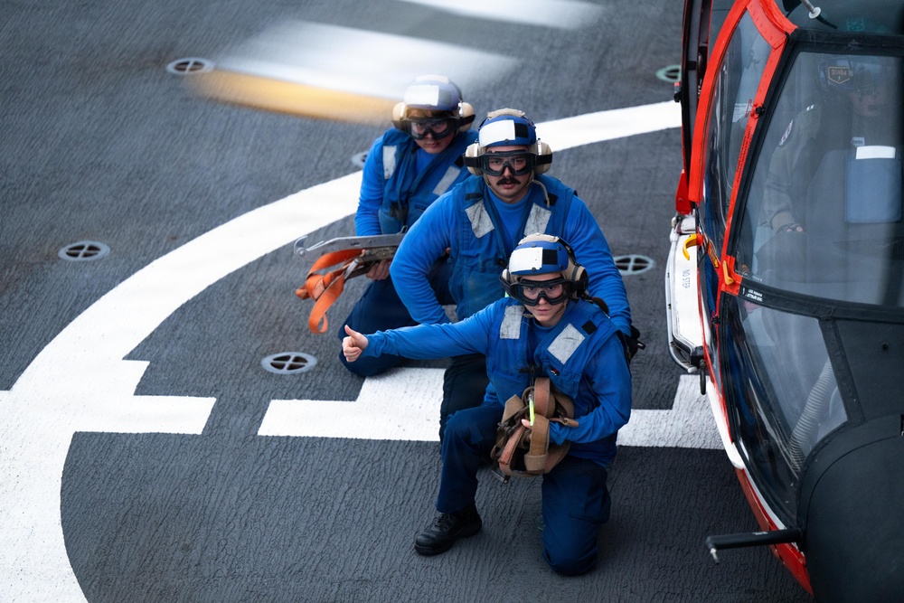 Three personnel in blue flight deck uniforms are positioned on a helicopter landing pad next to a red and white helicopter. They are wearing helmets, ear protection, and safety goggles, indicating an active flight deck operation. The foreground individual is giving a thumbs-up gesture, suggesting a readiness or all-clear signal.