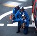 Three personnel in blue flight deck uniforms are positioned on a helicopter landing pad next to a red and white helicopter. They are wearing helmets, ear protection, and safety goggles, indicating an active flight deck operation. The foreground individual is giving a thumbs-up gesture, suggesting a readiness or all-clear signal.