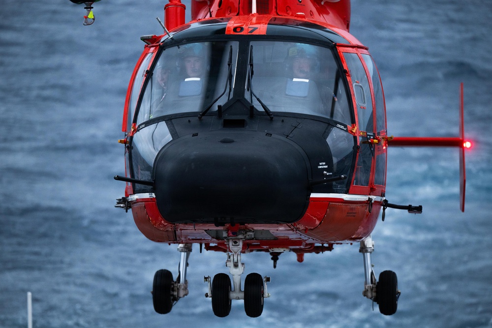 A red helicopter, numbered '07', is captured in a front-facing close-up shot hovering over open ocean water. Two pilots are visible through the large cockpit windshield. The helicopter appears to be a search and rescue or coast guard variant, equipped with a nose-mounted sensor pod and retractable landing gear.