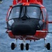 A red helicopter, numbered '07', is captured in a front-facing close-up shot hovering over open ocean water. Two pilots are visible through the large cockpit windshield. The helicopter appears to be a search and rescue or coast guard variant, equipped with a nose-mounted sensor pod and retractable landing gear.
