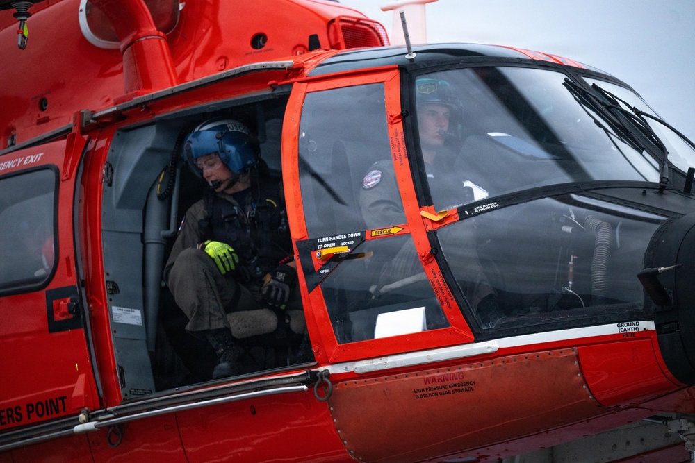 A close-up photograph of a red rescue helicopter, likely a U.S. Coast Guard aircraft, showing two crew members inside the cockpit and cabin area. One crew member in the cabin is wearing a blue flight helmet and bright yellow-green gloves, while a pilot is visible through the cockpit windshield. The helicopter displays safety markings including 'RESCUE,' 'EMERGENCY EXIT,' and warnings about flotation gear storage.
