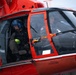 A close-up photograph of a red rescue helicopter, likely a U.S. Coast Guard aircraft, showing two crew members inside the cockpit and cabin area. One crew member in the cabin is wearing a blue flight helmet and bright yellow-green gloves, while a pilot is visible through the cockpit windshield. The helicopter displays safety markings including 'RESCUE,' 'EMERGENCY EXIT,' and warnings about flotation gear storage.