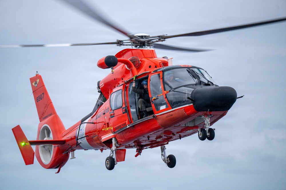 A bright orange-red U.S. Coast Guard MH-65 Dolphin helicopter (tail number 6507) is captured in flight against an overcast sky. The side door is open with a crew member visible, suggesting a search and rescue or training operation. The image is a high-quality, close-range aerial photograph showing significant detail of the aircraft.