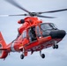 A bright orange-red U.S. Coast Guard MH-65 Dolphin helicopter (tail number 6507) is captured in flight against an overcast sky. The side door is open with a crew member visible, suggesting a search and rescue or training operation. The image is a high-quality, close-range aerial photograph showing significant detail of the aircraft.