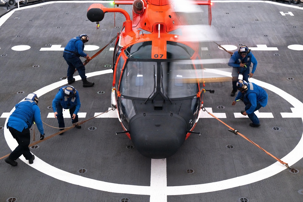 An orange and black helicopter marked '07' is being secured on a ship's flight deck helipad by a crew of five personnel dressed in blue uniforms. The crew members are using orange tie-down straps to restrain the helicopter while its rotor blades are still spinning, as indicated by the motion blur. This appears to be a U.S. Coast Guard helicopter landing or securing operation aboard a vessel.