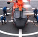 An orange and black helicopter marked '07' is being secured on a ship's flight deck helipad by a crew of five personnel dressed in blue uniforms. The crew members are using orange tie-down straps to restrain the helicopter while its rotor blades are still spinning, as indicated by the motion blur. This appears to be a U.S. Coast Guard helicopter landing or securing operation aboard a vessel.