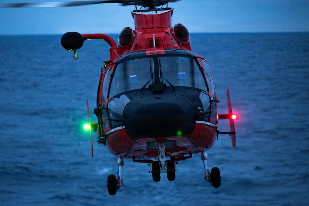 A red search-and-rescue helicopter is captured in a head-on view hovering over open ocean water at dusk or dawn. The aircraft displays active navigation lights — green on the left side and red on the right — indicating it is airborne and in operation. The number '07' is visible on the fuselage, suggesting it is a numbered unit, consistent with U.S. Coast Guard MH-65 Dolphin helicopter markings.