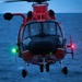 A red search-and-rescue helicopter is captured in a head-on view hovering over open ocean water at dusk or dawn. The aircraft displays active navigation lights — green on the left side and red on the right — indicating it is airborne and in operation. The number '07' is visible on the fuselage, suggesting it is a numbered unit, consistent with U.S. Coast Guard MH-65 Dolphin helicopter markings.