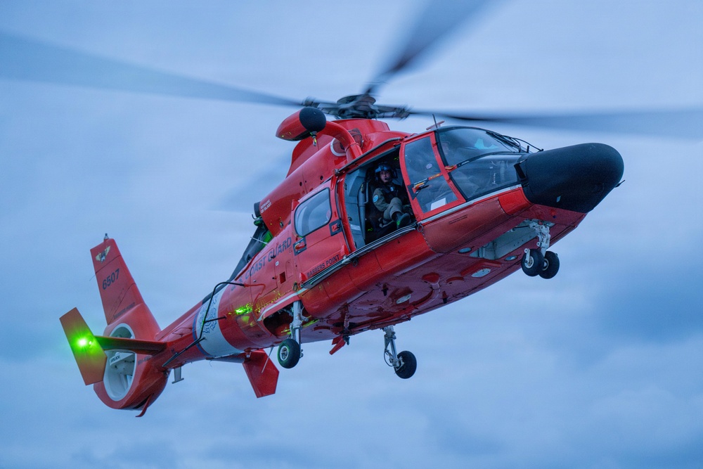 A bright red U.S. Coast Guard helicopter, identified by the number 6907 on its tail, is captured in flight against an overcast blue-grey sky. The helicopter appears to be a Eurocopter/Airbus HH-65 Dolphin, a common Coast Guard search-and-rescue aircraft. At least one crew member is visible through the open side door, wearing flight gear and a helmet.