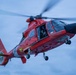 A bright red U.S. Coast Guard helicopter, identified by the number 6907 on its tail, is captured in flight against an overcast blue-grey sky. The helicopter appears to be a Eurocopter/Airbus HH-65 Dolphin, a common Coast Guard search-and-rescue aircraft. At least one crew member is visible through the open side door, wearing flight gear and a helmet.