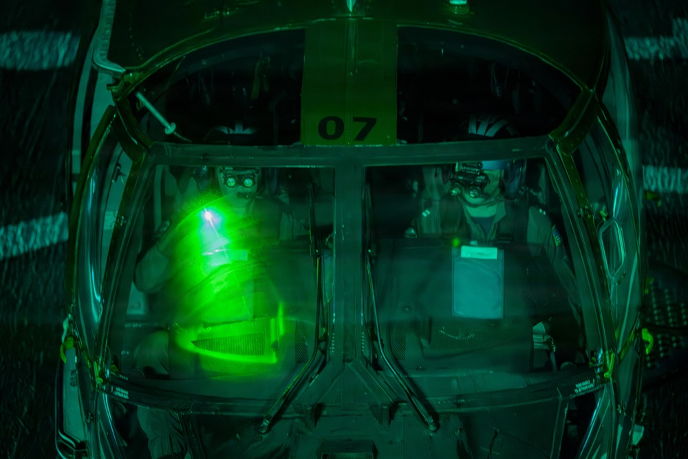 A top-down view into a military aircraft cockpit, likely a helicopter, illuminated by intense green light consistent with night vision equipment or green cockpit lighting. Two crew members are visible through the canopy glass, with the number '07' stenciled on the airframe. The image was taken at night, with the green glow dominating the interior.