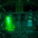 A top-down view into a military aircraft cockpit, likely a helicopter, illuminated by intense green light consistent with night vision equipment or green cockpit lighting. Two crew members are visible through the canopy glass, with the number '07' stenciled on the airframe. The image was taken at night, with the green glow dominating the interior.
