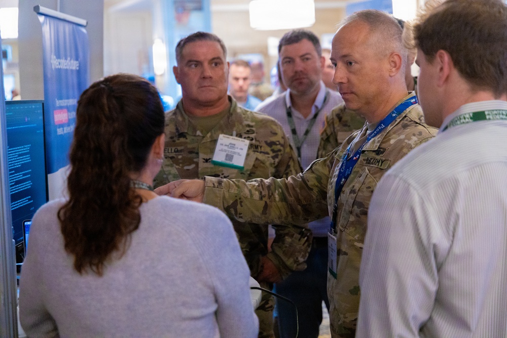 A group of people are gathered at what appears to be a technology conference or trade show booth. Two individuals in U.S. Army camouflage uniforms are engaged in conversation with a civilian presenter near a display screen. The setting suggests a military-industry technology demonstration or briefing event.
