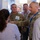 A group of people are gathered at what appears to be a technology conference or trade show booth. Two individuals in U.S. Army camouflage uniforms are engaged in conversation with a civilian presenter near a display screen. The setting suggests a military-industry technology demonstration or briefing event.