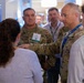 A group of people are gathered at what appears to be a technology conference or trade show booth. Two individuals in U.S. Army camouflage uniforms are engaged in conversation with a civilian presenter near a display screen. The setting suggests a military-industry technology demonstration or briefing event.
