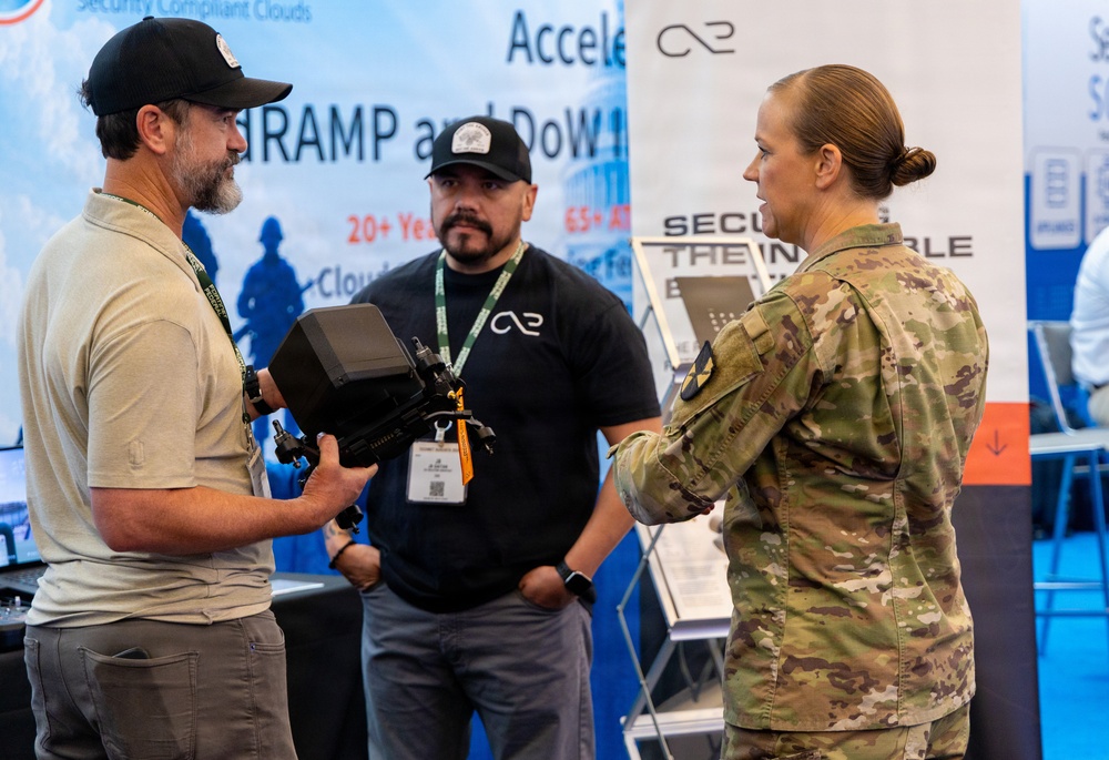 A military service member in OCP (Operational Camouflage Pattern) uniform is engaged in a discussion with two civilian vendors at what appears to be a defense or technology trade show/expo. One civilian is holding a piece of electronic equipment, likely a military or surveillance device, while the other stands nearby. The background displays vendor banners referencing FedRAMP, DoD, and cloud security compliance services.