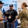 A military service member in OCP (Operational Camouflage Pattern) uniform is engaged in a discussion with two civilian vendors at what appears to be a defense or technology trade show/expo. One civilian is holding a piece of electronic equipment, likely a military or surveillance device, while the other stands nearby. The background displays vendor banners referencing FedRAMP, DoD, and cloud security compliance services.