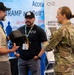 A military service member in OCP (Operational Camouflage Pattern) uniform is engaged in a discussion with two civilian vendors at what appears to be a defense or technology trade show/expo. One civilian is holding a piece of electronic equipment, likely a military or surveillance device, while the other stands nearby. The background displays vendor banners referencing FedRAMP, DoD, and cloud security compliance services.
