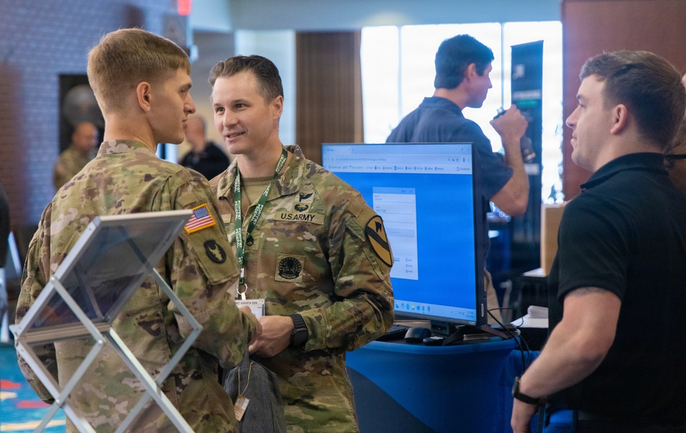Two U.S. Army soldiers in OCP (Operational Camouflage Pattern) uniforms are engaged in conversation with a civilian at what appears to be a military technology expo or conference. A computer monitor displaying a software interface is visible on a blue barrel-style table. The setting suggests a vendor demonstration or technology showcase event.