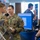 Two U.S. Army soldiers in OCP (Operational Camouflage Pattern) uniforms are engaged in conversation with a civilian at what appears to be a military technology expo or conference. A computer monitor displaying a software interface is visible on a blue barrel-style table. The setting suggests a vendor demonstration or technology showcase event.