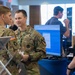 Two U.S. Army soldiers in OCP (Operational Camouflage Pattern) uniforms are engaged in conversation with a civilian at what appears to be a military technology expo or conference. A computer monitor displaying a software interface is visible on a blue barrel-style table. The setting suggests a vendor demonstration or technology showcase event.