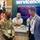 The image shows a trade show or conference exhibit floor featuring a ServiceNow booth. A U.S. Army soldier in camouflage uniform is engaged in a cheerful conversation with a man in a light blue blazer, both wearing green lanyards with badges. A third person sits behind a counter in the background, and a Cisco Partner booth is partially visible to the left.